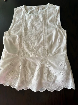 Vit broderad blus med spetsdetaljer - Söt vit blus i bomull med broderade detaljer och spets längs framsidan. Ärmlös modell med rund halsringning och vågig kant nedtill. 