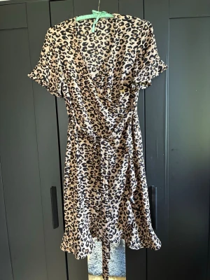 Leopardmönstrad omlottklänning - Säljer en kort leopardmönstrad omlottklänning med volangkant och korta ärmar. Klänningen har en beige bas med svarta och bruna leopardfläckar samt ett knytband i midjan. Perfekt för dig som vill sticka ut med ett djurmönster!