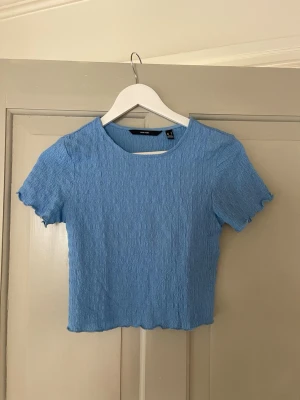Blå croppad topp från Vero Moda - Säljer en ljusblå croppad topp från Vero Moda. Toppen har korta ärmar, rund halsringning och är gjord i ett stretchigt, strukturerat material som ger en cool effekt. Perfekt till jeans eller kjol för en fräsch look. Den är som ny!
