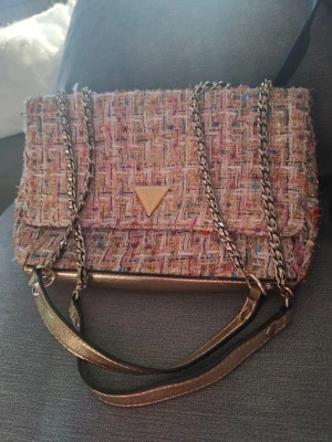 Tweed handväska med gulddetaljer Guess - Snygg handväska från Guess i flerfärgad tweed med guldfärgade metalldetaljer och kedjeremmar. Väskan har ett triangelformat Guess-emblem på framsidan och insidan är fodrad med logotypmönstrat tyg samt flera fack för småsaker.