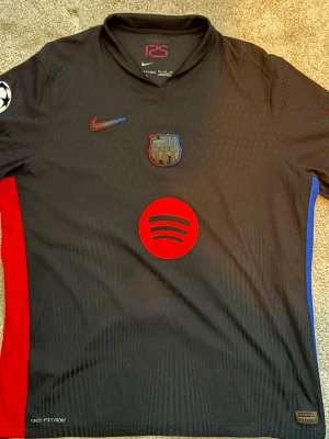 Svart FC Barcelona matchtröja Nike - Snygg svart FC Barcelona matchtröja från Nike med röda och blå detaljer på sidorna. Tröjan har klubbmärke, Nike-logga och Spotify som sponsor framtill samt Lamine Yamal och nummer 19 på ryggen. Tillverkad i lätt funktionsmaterial, perfekt för fotboll.