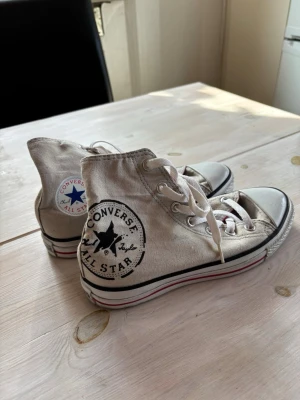 Converse All Star high tops - Klassiska Converse All Star high tops med svart logga på sidan och vit gummisula med röd och svart rand. Skorna har vita snören och metallhål, samt röd insida. Tillverkade i canvas för en tidlös och cool look.