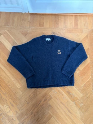 Mörkblå stickad tröja Gina Tricot - Mysig mörkblå stickad tröja från Gina Tricot i ullblandning. Tröjan har rund halsringning, ribbade muddar och en liten broderad nalle på bröstet som detalj. Perfekt för kalla dagar och enkel att matcha med jeans eller kjol.