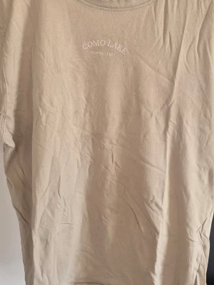 Beige t-shirt med Como Lake-tryck - Säljer en beige t-shirt med trycket 'Como Lake North Italy' på bröstet. T-shirten har rund hals och korta ärmar. Perfekt för dig som gillar enkel och stilren design.