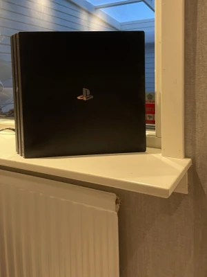 PlayStation 4 Pro spelkonsol svart - Säljer en svart PlayStation 4 Pro med den klassiska PlayStation-loggan på ovansidan. Konsolen har en stilren, fyrkantig design med tre lager och är tillverkad i plast. Perfekt för gaming och streaming.