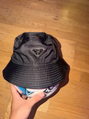 Svart bucket hat från Prada - Snygg svart bucket hat från Prada i glansigt nylonmaterial. Klassisk form med bred skärm och ikoniska metalltriangeln med logga framtill. Perfekt accessoar för en trendig streetwear-look.