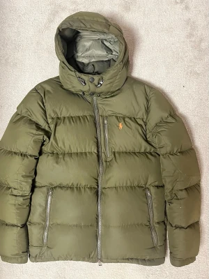 Olivgrön dunjacka från Polo Ralph Lauren - Olivgrön dunjacka med huva från Polo Ralph Lauren. Jackan har dragkedja framtill, tre fickor med dragkedja och ikonisk logga på bröstet. Perfekt för kalla dagar med sin tjocka vaddering och stilrena look.