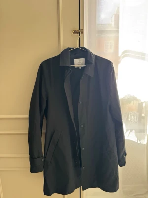 Svart rock från Bläck, storlek 48 - Stilren svart rock från Bläck med klassisk krage och knäppning framtill. Skandinavisk design med raka linjer och diskreta fickor. Perfekt för dig som gillar minimalistisk och tidlös stil.