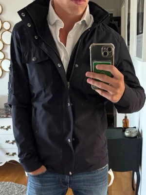 Rock&blue field jacka - Snygg svart vindjacka med hög krage och dragkedja framtill. Jackan har två bröstfickor med lock och knappar samt en ficka på ärmen. Tillverkad i ett lätt och vindtätt material, perfekt för blåsiga dagar. Passar till jeans och skjorta för en clean look.
