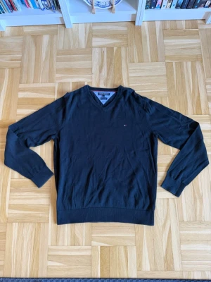 Svart V-ringad tröja Tommy Hilfiger - Klassisk svart V-ringad tröja från Tommy Hilfiger med diskret logga på bröstet. Tröjan har långa ärmar, ribbade muddar och är tillverkad i ett mjukt, tunt material som passar perfekt till lager på lager.