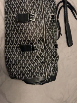Svart weekendbag med mönster - Snygg svart weekendbag med vitt grafiskt mönster och silverfärgade spännen. Väskan har dubbla handtag och justerbar axelrem med vadderad axelkudde. Materialet ser ut att vara konstläder med vita sömmar för extra detalj. Perfekt för resor eller gymmet. 