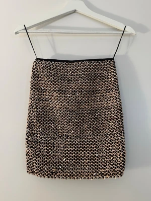 Glittrig kortkjol med paljetter - Säljer en glittrig kortkjol med tättsydda paljetter i svart och beige. Kjolen har en rak passform och är perfekt för dig som vill sticka ut. Materialet känns syntetiskt och kjolen har en cool, trendig vibe.