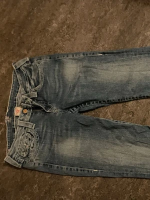 Blå jeans med kontrastsömmar - Säljer ett par blå jeans med slitningar och kontrastsömmar. Jeansen har fem fickor, knappgylf och detaljerade bakfickor med lock och knapp. Materialet är klassisk denim i bomull och passformen är rak.
