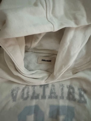 Vit hoodie från Zadig & Voltaire - Snygg vit hoodie från Zadig & Voltaire med trycket 'VOLTAIRE 97' i ljusgrått framtill. Hoodien har klassisk huva och magficka, samt ribbade muddar vid ärmslut och nederkant. Perfekt för en avslappnad och trendig look.