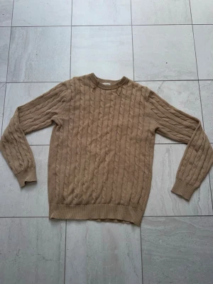 Beige John Henric tröja - Snygg beige kabelstickad tröja från John Henric. Tröjan har rund halsringning, ribbade muddar och nederkant samt ett klassiskt flätmönster. Perfekt för dig som gillar stilrena och tidlösa plagg i garderoben.
