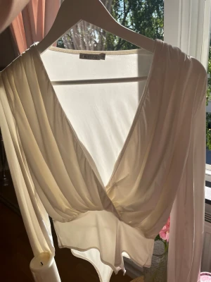 Vit långärmad body med omlott - Säljer en elegant vit body med lång ärm och omlottdesign framtill. Djup V-ringning och mjukt, stretchigt material som ger en snygg siluett. Perfekt att styla till både jeans och kjol för en trendig look.