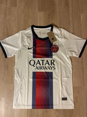 PSG matchtröja vit DOUE - Snygg vit PSG matchtröja med vertikal röd och blå rand på framsidan. Tröjan har svarta detaljer vid krage och ärmslut samt PSG-logga och Qatar Airways-tryck på bröstet. Perfekt för fotbollsfans som vill sticka ut på planen eller läktaren. HELT NY!