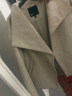Beige kappa med bred krage från lager 157 - Stilren beige kappa med bred krage och dubbelknäppning framtill. Kortare modell med raka fickor och mjukt ull-liknande material. Perfekt för dig som vill ha en trendig och enkel look.