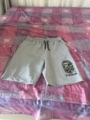 Grå shorts med camo-tryck från BAPE - Snygga grå shorts med elastisk midja och svart snörning. På vänster ben finns ett stort kamouflagefärgat BAPE-tryck och texten 'BY BATHING APE'. Perfekta för sommaren och chill dagar.