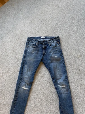 Replay jeans - Säljer ett par blå  jeans med slitningar och distressed detaljer på både fram- och baksida. Jeansen har fem fickor, knappgylf och klassisk passform. Perfekta för en avslappnad och trendig outfit. De är replay och sitter rätt så slimmat. Hör av dig vid frågor! De är 29 i waist och 32 i längd, men passar för mindre