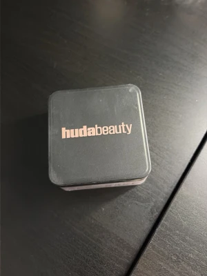 Huda Beauty Easy Bake Loose Powder - Säljer ett Huda Beauty Easy Bake löspuder i en fyrkantig burk med svart lock och rosa text. Pudret har en ljusrosa ton och kommer i en genomskinlig plastförpackning. Perfekt för att fixera foundation och ge en matt finish.