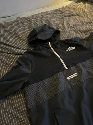 Svart vindjacka från The North Face - Snygg svart vindjacka från The North Face med halv dragkedja framtill och huva. Jackan har ett rutmönstrat parti över magen och vita logotyper på bröstet och magen. Tillverkad i lätt och tåligt material, perfekt för blåsiga dagar. Bra skick men finns spår av användning, nån enstaka fläck finns men går nog o tvätta bort