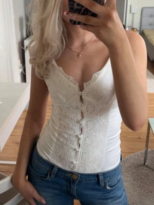 Vit spetsbody med knappar - Säljer en vit body med spetsdetaljer och knappar framtill. Djup V-ringning och smala axelband ger en feminin look🤩💗