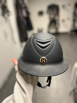 Svart ridhjälm med gulddetaljer från Onek - Snygg svart ridhjälm med ventilerande meshdetaljer framtill och guldfärgade detaljer från Onek. Hjälmen har en klassisk form med skärm och justerbart hakband. Den har lite små repor där av priset. Reporna syns knappt. Har inte ramlat av med den den. Perfekt för dig som vill ha både stil och funktion i stallet. 