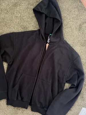 Skims Svart zip hoodie med huva - En klassisk svart hoodie med dragkedja framtill och stor huva. Hoodien har två rymliga fickor på magen och ribbade muddar vid ärmslut och nederkant. Perfekt för en avslappnad och stilren look. Materialet känns mjukt och bekvämt.