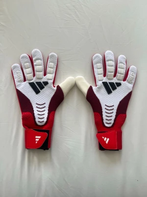 Adidas predator målvaktshandskar - Snygga målvaktshandskar i rött och vitt med svarta detaljer. Handskarna har greppvänlig insida och vadderade fingrar för extra skydd. Justerbar kardborre vid handleden för bästa passform. Perfekta för dig som älskar fotboll! Helt nya med plast och storlek 9