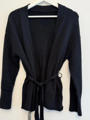Cardigan - Klassisk svart cardigan i öppen modell. Har ett löst knytband i midjan som ger en fin passform, sänkta axelsömmar och ribbstickad kant vid armsluten. En mångsidig basfavorit i garderoben.