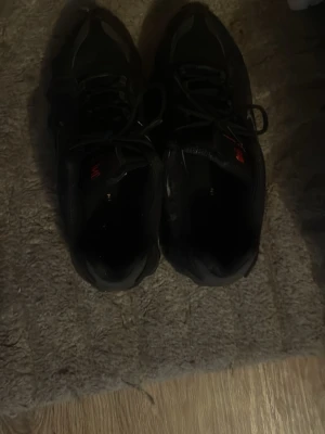 Svarta sneakers med reflexdetaljer - Säljer ett par svarta sneakers med coola reflexdetaljer och snörning. Skorna har en sportig look med robust sula och diskreta röda inslag på plösen. Perfekta för dig som gillar streetwear och vill ha något stilrent till vardags.