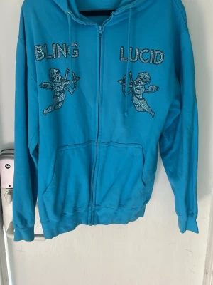 Blå hoodie med bling och änglar - Säljer en blå zip-hoodie med huva och fickor framtill. På bröstet står det 'BLING' och 'LUCID' i glittriga stenar, samt två änglamotiv i samma stil. Perfekt för dig som gillar streetwear och vill sticka ut med coola detaljer.