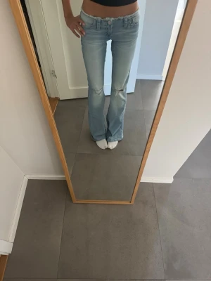 Ljusblå bootcut jeans med rå kant - Säljer ett par ljusblå bootcut jeans med rå, fransad kant nedtill. Jeansen har låg midja, klassiska fem fickor och slitningar vid knäna för en trendig look. Perfekta för dig som gillar en avslappnad och cool stil.