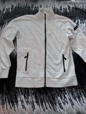   sail racing Vit ziptröja med svarta detaljer - Snygg vit ziptröja med hög krage och svarta dragkedjor på både framficka och mitt. Tröjan har ribbade muddar vid ärmslut och nederkant samt en diskret svart patch på ärmen. Perfekt för en clean och sportig look.