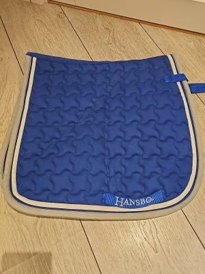 Blått schabrak från Hansbo - Säljer ett blått schabrak från Hansbo med beige kantband och quiltat mönster. Perfekt till hästutrustning och har praktiska fästen. Passar till sadel och ger en snygg look i stallet. Storlek ponny och bara andvänd 1 gång!