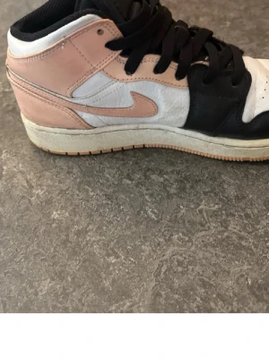 Svarta, vita och rosa sneakers - Snygga sneakers i svart, vitt och ljusrosa med klassisk Nike swoosh på sidan. Skorna har svarta skosnören och en vit sula med beige yttersula. Modellen är mid och har en ikonisk design med paneler i olika färger och material i läder.