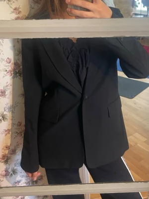 Svart klassisk kavaj med fickor - Stilren svart kavaj med enkelknäppning och två framfickor. Kavajen har en klassisk passform med smala slag och knappar vid ärmsluten. Perfekt att matcha med både byxor och kjol för en snygg look.