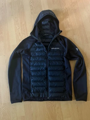 Peak performance jacka  - Snygg svart pufferjacka från Peak Performance med quiltad framsida och rygg, samt släta ärmar och sidor. Jackan har huva och dragkedja framtill. Perfekt för kalla vinterdagar och har en sportig look. Använd ett fåtal gånger. Hör av dig om du undrar något!