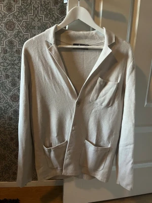 Beige cardigan med fickor - Stilren beige cardigan med tre framfickor och knappstängning. Tröjan har klassisk krage och är tillverkad i ett mjukt, tunt stickat material. Perfekt för lager-på-lager och enkel att matcha med olika outfits.