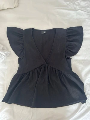 Svart blus med volangärmar - Säljer en svart blus med djup v-ringning och stora volangärmar. Toppen har en lätt utsvängd passform och är gjord i ett ribbat material som ger en snygg struktur. Perfekt att styla med jeans eller kjol för en trendig look.