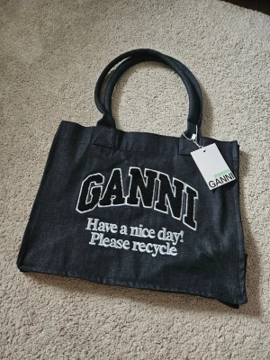 Tote Bag från Ganni - Cool svart shoppingväska från Ganni i slitstarkt tyg med vita broderade bokstäver och texten 'Have a nice day! Please recycle' på framsidan. Väskan har dubbla handtag och en enkel, rymlig design. Perfekt för att bära allt du behöver till skolan eller stan.