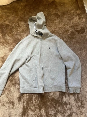 Grå zip hoodie med broderad loggas  - Säljer en stilren grå hoodie med dragkedja och huva. Tröjan har två fickor framtill och en liten broderad logga på bröstet. Perfekt för chill dagar eller när du vill ha en enkel men snygg look. Materialet känns mjukt och bekvämt.