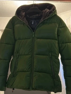 Save The Duck Jacka - Snygg grön pufferjacka med huva och dragkedja framtill. Jackan har två sidofickor med dragkedja och quiltad design som ger en riktigt puffig look. Perfekt för kalla dagar och ger en trendig streetwear-vibe.
