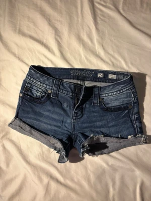 Blå jeansshorts från Miss Me - Säljer ett par blå jeansshorts från Miss Me med råa kanter och mycket detaljer. Shortsen har knappgylf och dekorativa sömmar samt ficklock bak. Perfekta för sommaren och ger en avslappnad vibe.