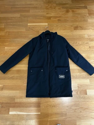 Svart regnjacka från Vans med huva - Snygg svart regnjacka från Vans med huva och dragkedja framtill. Jackan har två stora fickor med tryckknappar och en tydlig Vans-logga på ena fickan. Lång modell som skyddar bra mot regn, perfekt för blöta dagar. Använd ca 3 gånger , som ny. 