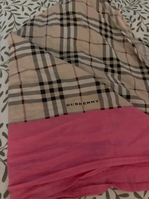 Burberry  - The images show a scarf with the classic Burberry plaid pattern in beige, black, and red, featuring a prominent pink border. The word 'BURBERRY' is visible on the scarf, indicating the brand. The scarf is laid out on a bed with a leafy patterned bedspread.