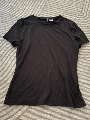 Svart basic t-shirt  - En enkel svart t-shirt med rund hals och korta ärmar. Tillverkad i tunn bomull som känns lätt och skön mot huden. Perfekt att matcha med jeans eller kjol för en clean look.
