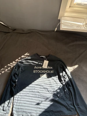 Marinblå långärmad topp Acne Studios - Svart långärmad topp från Acne Studios med tryck i vitt framtill där det står 'Acne Studios STOCKHOLM 1996'. Klassisk rund halsringning och mjukt bomullsmaterial. Perfekt för en clean och stilren look. Skriv om du har frågor innan köp!