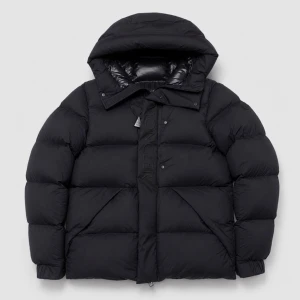 Svart pufferjacka från Moncler - Snygg svart pufferjacka från Moncler med stor huva och diskret logga på ärmen. Jackan har en boxig passform, dragkedja och tryckknappar framtill samt två stora fickor. Perfekt för kalla dagar när du vill hålla stilen och värmen. Modellen av jackan heter Madeira och är perfekt nu för vintern.storlek 3=M. Taggar kommer med